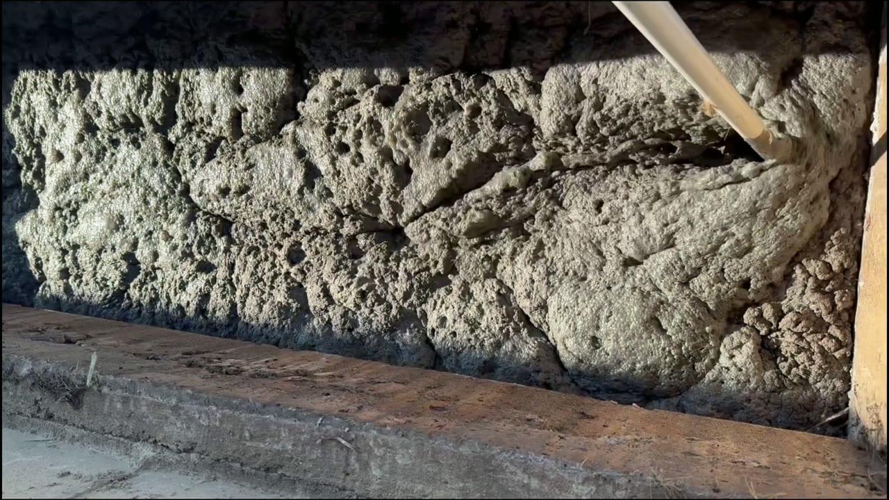 A manure pit photographed beneath the barn slats, its surface built up into a thick, hardened crust, with a treatment pipe reaching down through the gap.